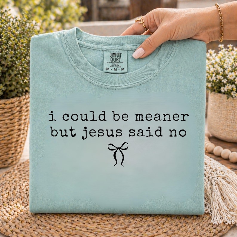 Op de afbeelding: Lichtblauw T-shirt met de zwarte tekst "i could be meaner but jesus said no" en een strik-afbeelding. Het T-shirt wordt vastgehouden door een hand met gouden sieraden.