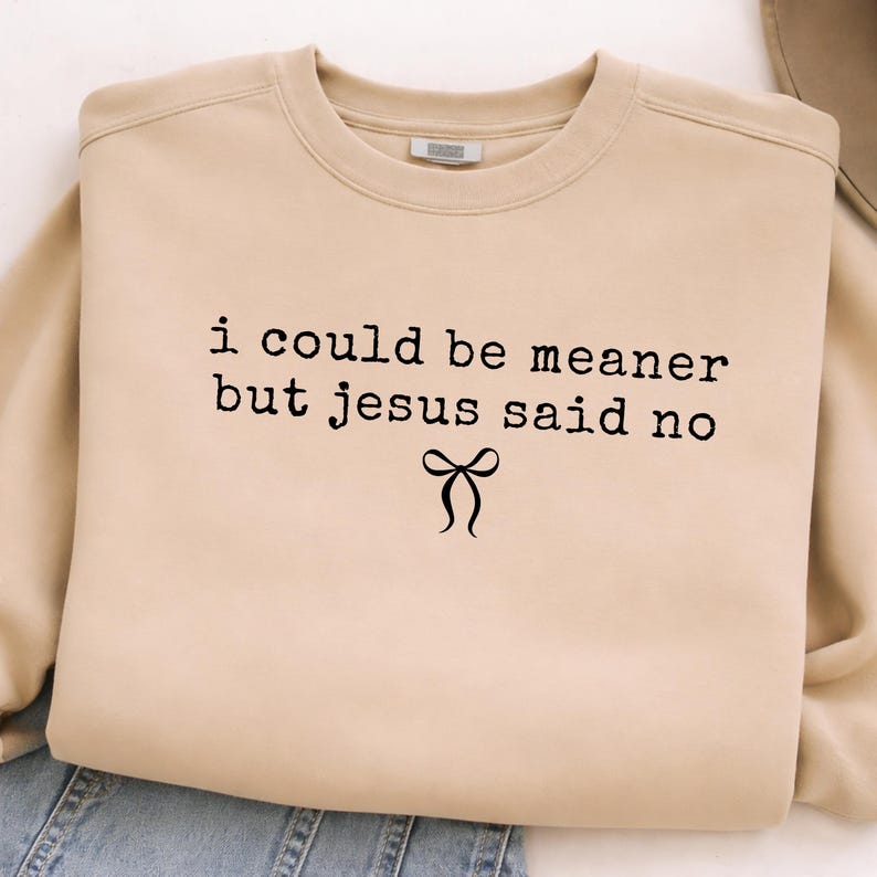 Op de afbeelding: Een beige sweatshirt met ronde hals en de zwarte tekst "i could be meaner but jesus said no", met een strik-afbeelding. De casual sweatshirt is gemaakt van zacht materiaal, geschikt voor dagelijks gebruik.