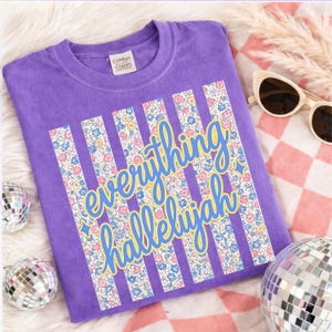 May include: A purple Comfort Colors t-shirt with the words "everything hallelujah" in a script font with a yellow outline. The text is set against vertical stripes filled with a floral pattern. The shirt is displayed with sunglasses and disco balls.