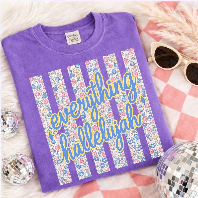 May include: A purple Comfort Colors t-shirt with the words "everything hallelujah" in a script font with a yellow outline. The text is set against vertical stripes filled with a floral pattern. The shirt is displayed with sunglasses and disco balls.
