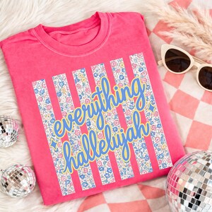 May include: A bright pink t-shirt with the words "everything hallelujah" in a blue and yellow script. The text is set against vertical stripes filled with a floral pattern. The shirt is displayed on a soft, white surface.