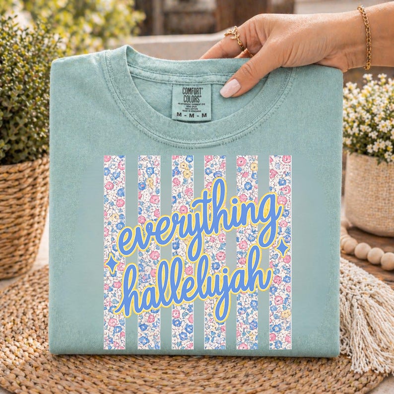 May include: A light teal Comfort Colors t-shirt with the words "everything hallelujah" in a cursive font. The design includes vertical floral stripes in pink, yellow, and blue. The shirt is folded and held by a person.
