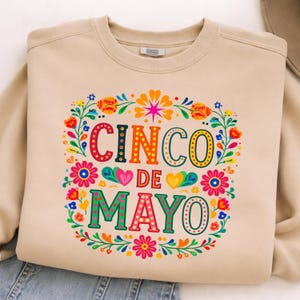 May include: A beige sweatshirt featuring a colourful floral design and the words "CINCO DE MAYO". The design includes flowers, hearts, and decorative elements, ideal for celebrating the holiday.