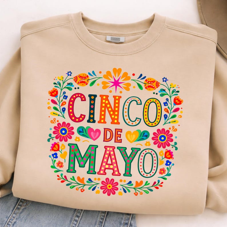 May include: A beige sweatshirt featuring a colourful floral design and the words "CINCO DE MAYO". The design includes flowers, hearts, and decorative elements, ideal for celebrating the holiday.