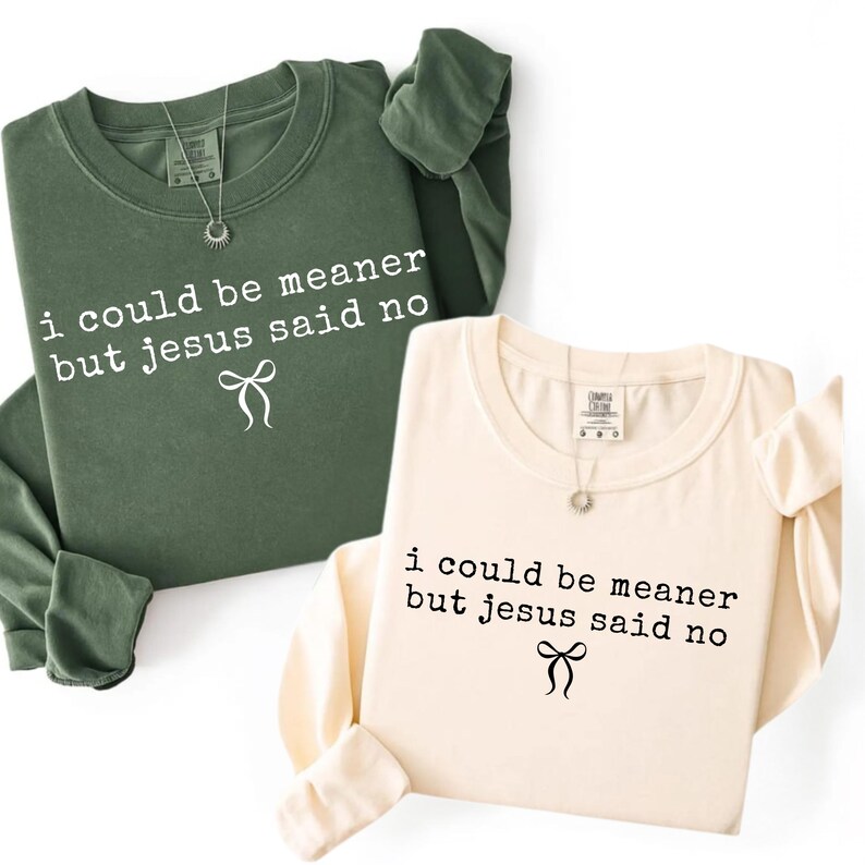 Op de afbeelding: Twee sweatshirts met lange mouwen, &eacute;&eacute;n olijfgroen en &eacute;&eacute;n cr&egrave;me, worden getoond. Elk shirt heeft de tekst "i could be meaner but jesus said no" in een zwarte letter, samen met een strik. Beide shirts hebben een ronde hals en een kleine hanger.