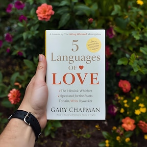 Puede incluir: Un libro titulado "5 Languages of Love" se sostiene frente a un fondo floral. La portada del libro es blanca con el título en letras grandes y un corazón rojo. También es visible el nombre del autor, Gary Chapman.