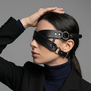 Puede incluir: Antifaz de cuero negro con anillos de metal plateado y cierres de hebilla. El antifaz tiene dos correas que se cruzan sobre los ojos y se aseguran alrededor de la cabeza. El diseño es simple y elegante.