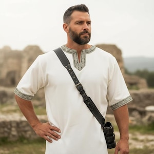 May include: A white tunic with short sleeves, featuring decorative trim around the neckline and sleeves. A black leather shoulder strap with a buckle and a small pouch is worn across the body. The tunic has a V-neck.