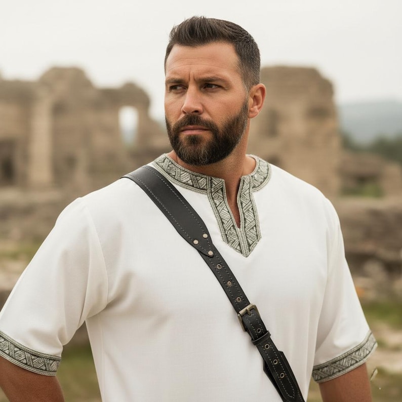 May include: A white tunic with decorative trim around the neckline and sleeves. A black leather strap with metal rivets and a buckle is worn across the chest. The person is outdoors with a blurred background of stone structures.