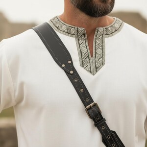 May include: Black leather shoulder strap with a metal buckle and rivets. The strap is worn over a white tunic with embroidered detailing around the neckline. The tunic has a V-neck design.