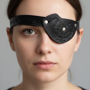 May include: Black leather eye patch featuring a compass rose design and silver rivets. The eye patch is attached to a black leather strap. The strap and eye patch have embossed decorative details. The eye patch is worn over one eye.