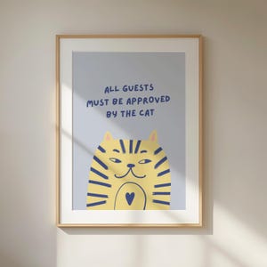 Könnte beinhalten: Gerahmter Kunstdruck mit einer Cartoon-Katze in Gelb und Blau. Der Text "ALL GUESTS MUST BE APPROVED BY THE CAT" steht darüber. Das Bild ist in einem Holzrahmen gefasst.