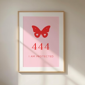 Könnte beinhalten: Ein gerahmter Wandkunstdruck mit einem hellrosa Hintergrund, einer roten Schmetterlingsgrafik oben, der Zahl 444 in der Mitte und dem Text I AM PROTECTED in roten Großbuchstaben unten, eingefasst in einen hellen Holzrahmen.