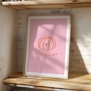 Könnte beinhalten: Ein gerahmter Kunstdruck mit rosa Hintergrund, einer orangefarbenen Kürbisillustration und einem Herz. Der Text "IN MY COZY ERA" befindet sich unter dem Kürbis. Der Druck wird auf einem Holzregal ausgestellt.