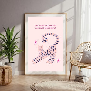 Könnte beinhalten: Ein gerahmter Kunstdruck mit einer rosa Katzenillustration mit blauen Wellenmustern und dem Text "WHY BE MOODY WHEN YOU CAN SHAKE YOUR BOOTY?" Zwei rosa Sterne sind ebenfalls im Design enthalten. Der Druck wird in einem Raum mit einer Pflanze und einem Korbstuhl präsentiert.
