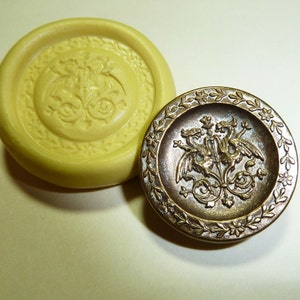 May include: A gold-toned metal button with a detailed design of three mythical creatures with wings and a crown. The button is surrounded by a decorative border of leaves and flowers.