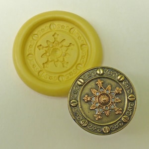 May include: A gold-colored metal button with a detailed snowflake design and a decorative border. The button is surrounded by a yellow silicone mold with a similar snowflake design.