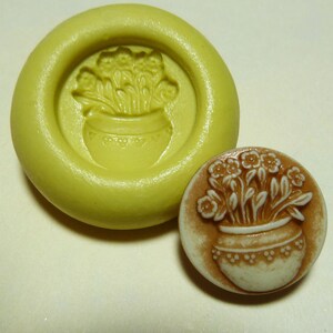 May include: A silicone mold for making a small, round, ceramic-style button with a raised design of a flower bouquet in a pot. The mold is yellow and the button is brown with white flowers.