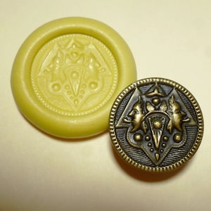 May include: A close-up of a mold for making a decorative button. The mold is yellow and has a design of two stylized faces with a central geometric shape. Next to the mold is a finished button made of brass with the same design.