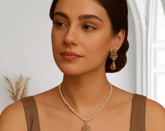 May include: A woman wearing a pearl necklace with a gold pendant and matching earrings. The pendant has a floral design with a teardrop-shaped charm. The jewelry is set against a brown top.