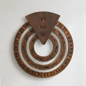 Puede incluir: Calendario perpetuo de madera en color marrón oscuro. Cuenta con anillos giratorios que muestran meses, días y fechas. La fecha actual es el 27 de septiembre, sábado. Una decoración de pared única.
