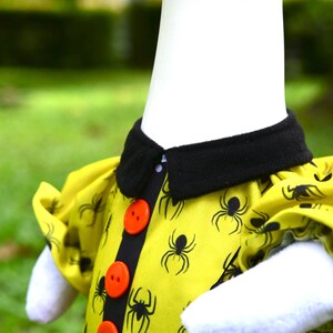 May include: A yellow shirt featuring a black spider pattern, a black collar, and three bright orange buttons down the center front. The garment has short puffed sleeves and is worn on a white mannequin torso against a blurred outdoor background.