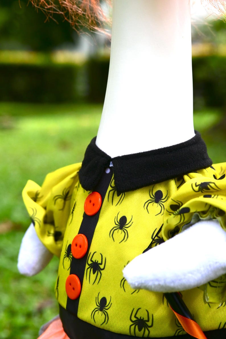 May include: A yellow shirt featuring a black spider pattern, a black collar, and three bright orange buttons down the center front. The garment has short puffed sleeves and is worn on a white mannequin torso against a blurred outdoor background.