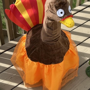 May include: A plush turkey costume with a brown head, large white eyes, and a yellow beak. The costume features a brown body, an orange tutu, and a colorful fan tail with red, orange, and yellow stripes. Perfect for Thanksgiving or Halloween.