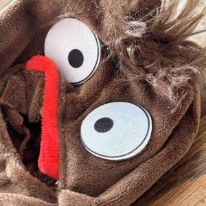May include: A brown plush costume hood with large white eyes, black pupils, and a red tongue. The hood has shaggy brown faux fur around the top. The hood is made of soft, plush material.