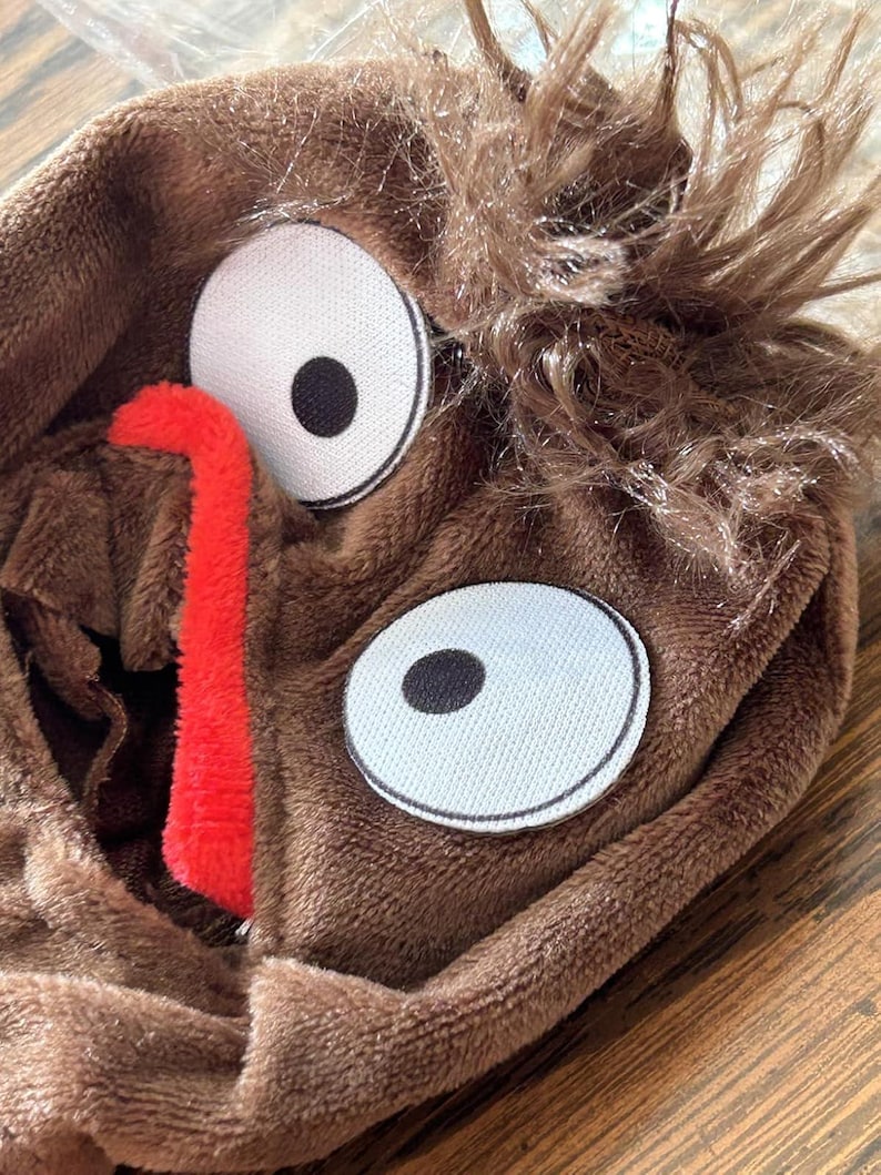 May include: A brown plush costume hood with large white eyes, black pupils, and a red tongue. The hood has shaggy brown faux fur around the top. The hood is made of soft, plush material.
