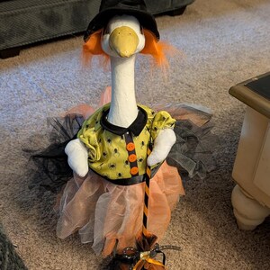 May include: A white goose figurine dressed as a witch, wearing a black pointed hat, orange hair, and a yellow shirt with a black spider pattern and orange buttons. The goose wears a layered orange and black tulle skirt and holds a miniature broom.