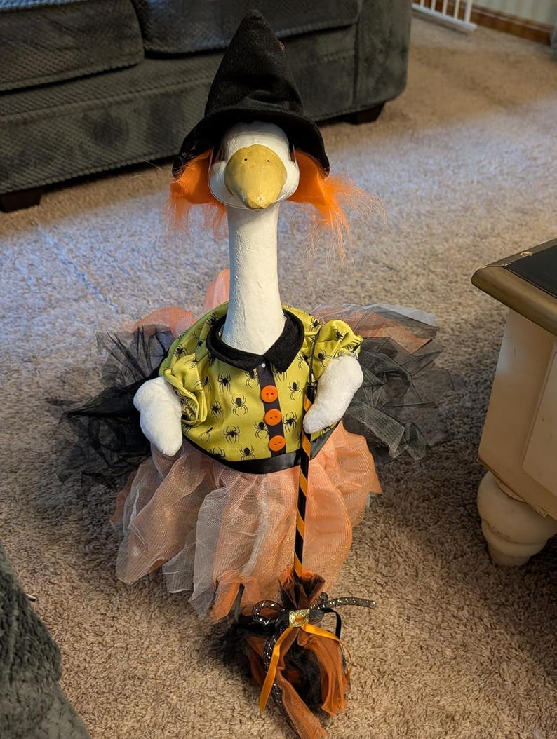 May include: A white goose figurine dressed as a witch, wearing a black pointed hat, orange hair, and a yellow shirt with a black spider pattern and orange buttons. The goose wears a layered orange and black tulle skirt and holds a miniature broom.