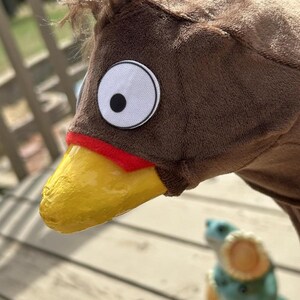 May include: A brown plush bird costume head with a yellow beak, red accent, and large white eye with a black pupil. The costume has a fluffy brown crest. A small, blurred figurine is in the background.