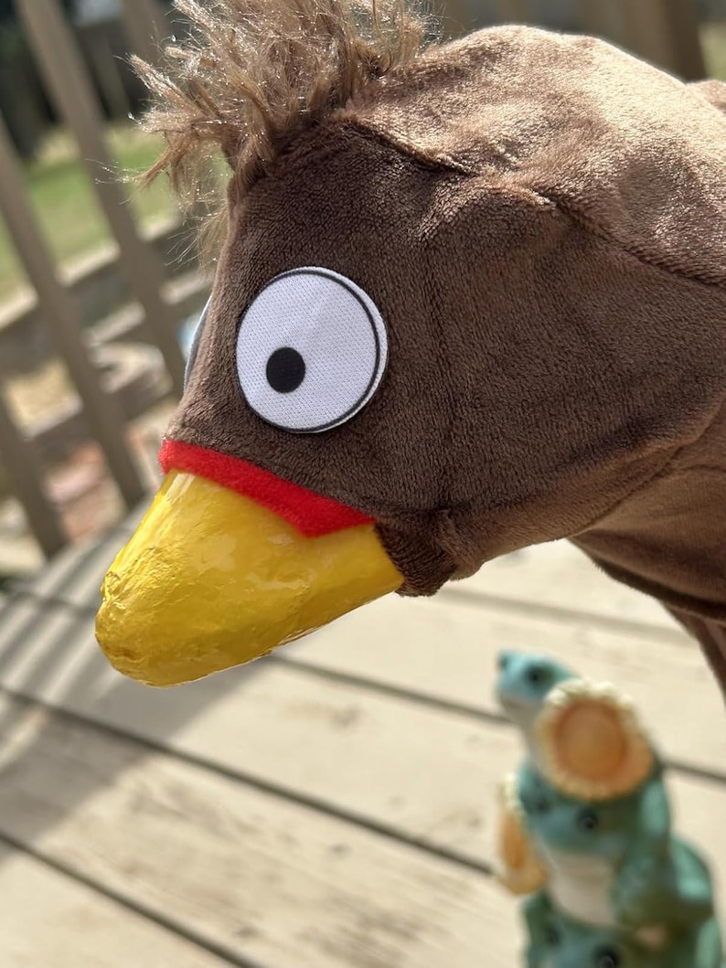 May include: A brown plush bird costume head with a yellow beak, red accent, and large white eye with a black pupil. The costume has a fluffy brown crest. A small, blurred figurine is in the background.