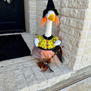 May include: A white goose figurine dressed as a witch, wearing a black pointed hat, bright orange hair, and a yellow shirt with a black spider pattern. The goose has a layered orange and black tulle skirt and holds a small broomstick accessory.