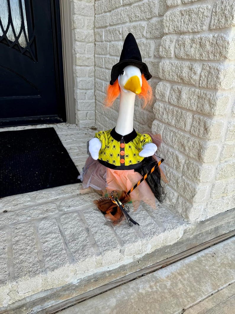 May include: A white goose figurine dressed as a witch, wearing a black pointed hat, bright orange hair, and a yellow shirt with a black spider pattern. The goose has a layered orange and black tulle skirt and holds a small broomstick accessory.