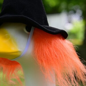 May include: A white goose figurine wearing a black felt witch hat and a bright orange synthetic wig. The hat is secured with a thin white elastic chin strap. The goose has a yellow beak and a smooth white neck, contrasting with the vibrant hair.