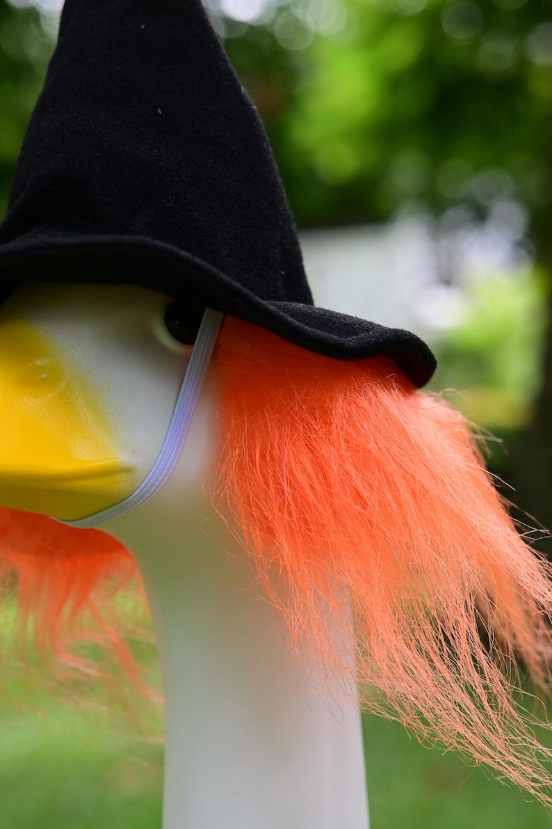 May include: A white goose figurine wearing a black felt witch hat and a bright orange synthetic wig. The hat is secured with a thin white elastic chin strap. The goose has a yellow beak and a smooth white neck, contrasting with the vibrant hair.