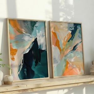 May include: Two framed abstract art prints with a wooden frame. The artwork features brushstrokes of teal, orange, white, and navy blue. The prints are displayed on a wooden shelf with books and plants.