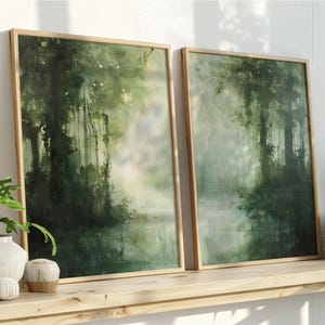 May include: Two framed landscape paintings with a forest scene in shades of green and white. The artwork is displayed on a wooden shelf with decorative items, including a potted plant and books. The frames are a light wood color.