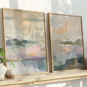 以下が含まれることがあります： 明るい木製フレームに入った2つの抽象的な風景画。 ピンク、青、緑などの柔らかなパステルカラーが特徴で、穏やかな雰囲気を作り出しています。 絵画は、装飾的な植物や本と一緒に木製の棚に飾られています。