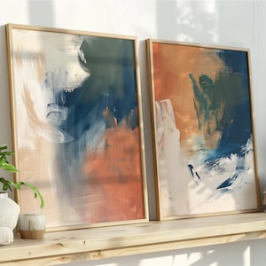 Könnte beinhalten: Zwei gerahmte abstrakte Kunstdrucke mit Holzrahmen. Das Kunstwerk zeigt Pinselstriche in Blau-, Orange- und Weißtönen. Die Drucke werden auf einem Holzregal mit dekorativen Gegenständen und Pflanzen ausgestellt.