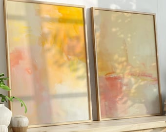 Puede incluir: Dos impresiones artísticas abstractas enmarcadas con tonos cálidos de amarillo, naranja y rosa se apoyan contra una pared blanca. Una estantería de madera debajo contiene un jarrón blanco con vegetación, pequeños objetos decorativos y una pila de libros.