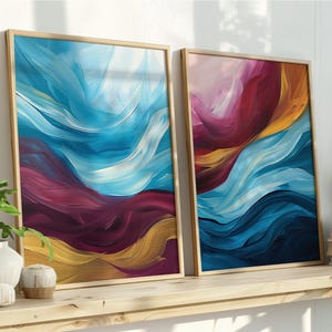 May include: Two framed abstract art prints with a gold border. The artwork features flowing brushstrokes in shades of blue, magenta, and gold. The prints are displayed on a wooden shelf with decorative items and plants.
