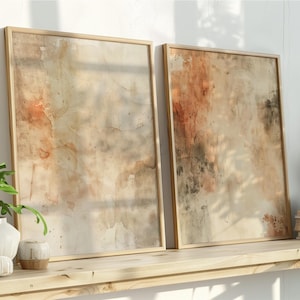 May include: Two framed abstract art prints with a watercolor effect in beige, brown, and black tones. The artwork is displayed on a light wood shelf with decorative plants and books. The frames are a light wood color.