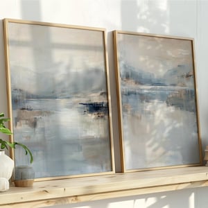 Puede incluir: Dos pinturas de paisajes abstractos enmarcadas con tonos neutros de azul, beige y gris. La obra de arte se exhibe en una estantería de madera clara con objetos decorativos y plantas. Las pinturas tienen una calidad serena, similar a la acuarela.