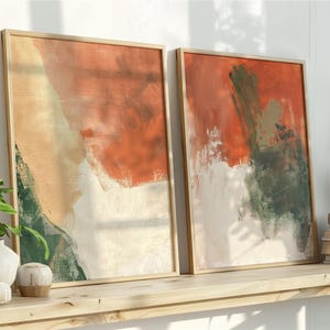 以下が含まれることがあります： 2つの額入り抽象芸術プリント。アートワークは、オレンジ、ベージュ、白、緑の色合いの大胆な筆致が特徴です。フレームは明るい木の色です。プリントは、装飾品や植物のある木製の棚に飾られています。