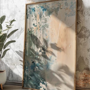 May include: Abstract art print in a wooden frame. The artwork features a textured design with shades of blue, beige, and white. The painting is leaning against a textured wall, with shadows cast across the surface.