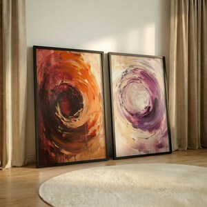 Pode incluir: Duas pinturas abstratas emolduradas com pinceladas giratórias. Uma apresenta tons quentes de laranja e vermelho, a outra roxos e cremes frios. Ambas estão em molduras pretas, encostadas a uma parede, com um tapete branco em primeiro plano.