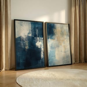 Könnte beinhalten: Zwei gerahmte abstrakte Kunstwerke. Das Kunstwerk zeigt eine Mischung aus tiefen Blautönen, Weiß und Gold, wodurch ein strukturierter, geschichteter Effekt entsteht. Die Rahmen sind schwarz, und die Kunst lehnt an einer Wand in der Nähe eines beigen Vorhangs und eines weißen Teppichs.